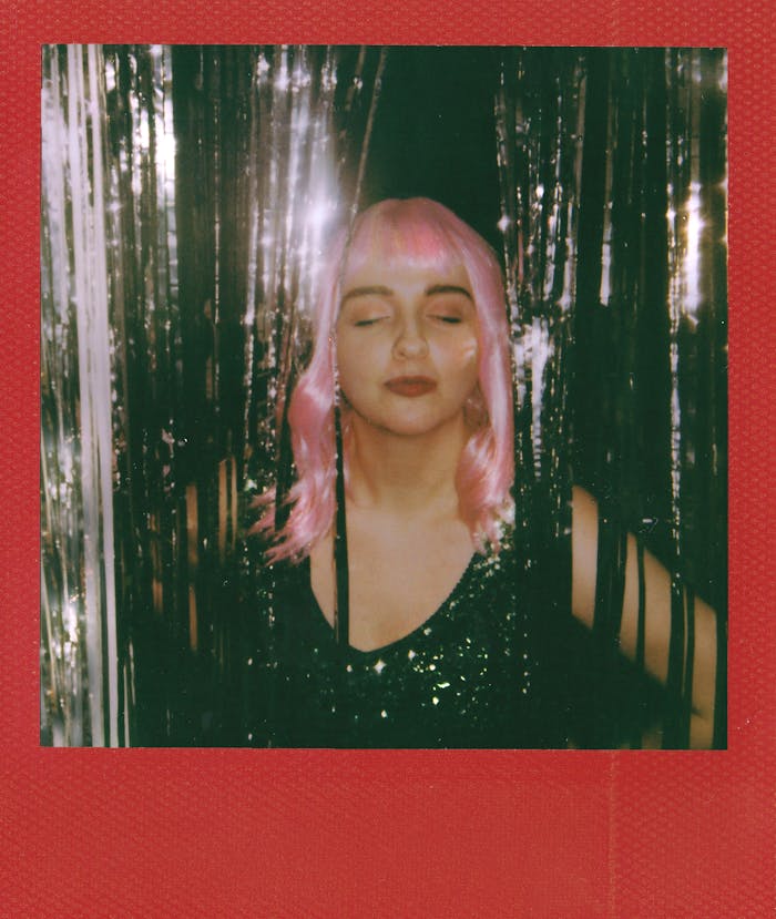 A woman with closed eyes wearing a pink wig and glitter dress, captured in a vibrant Polaroid photo.