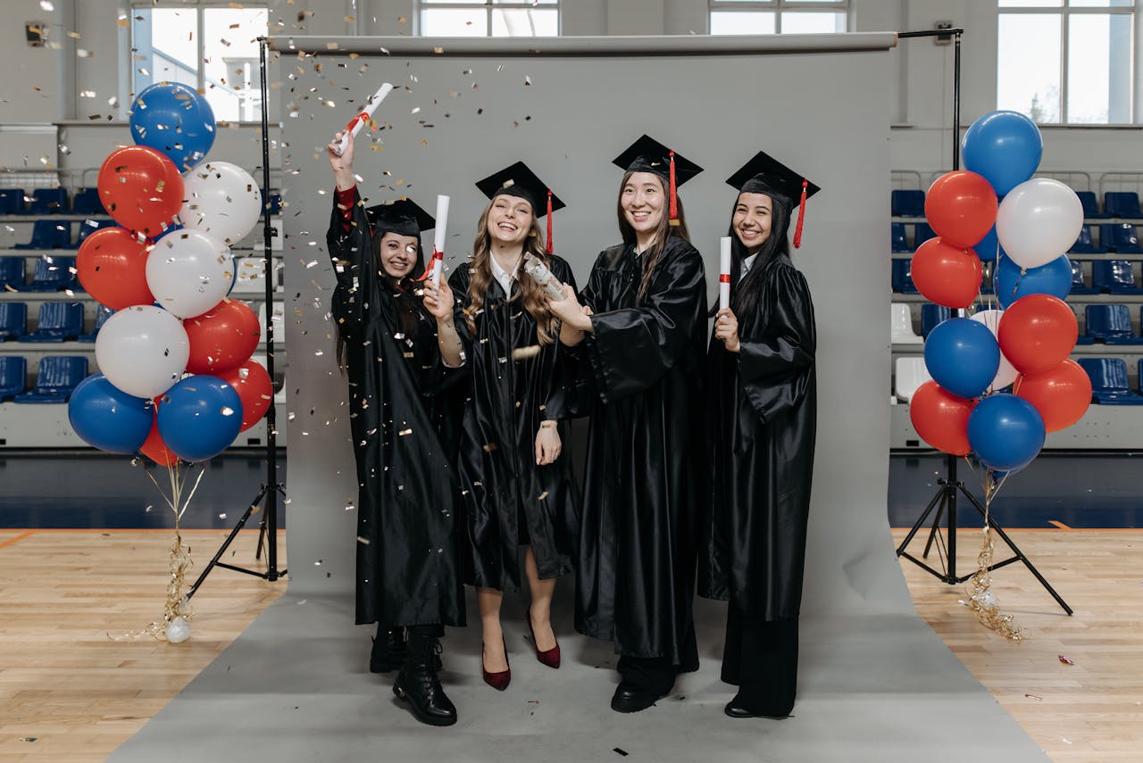 Four graduates celebrating with confetti and balloons in a gymnasium.